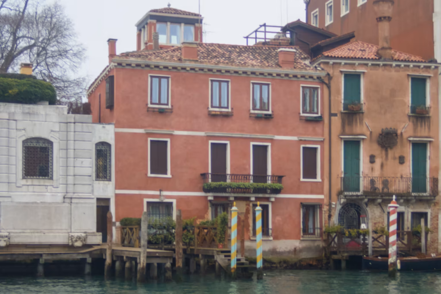 A fairly non-descript red three-storey building on a canal, with two turquose and yellow mooring posts.