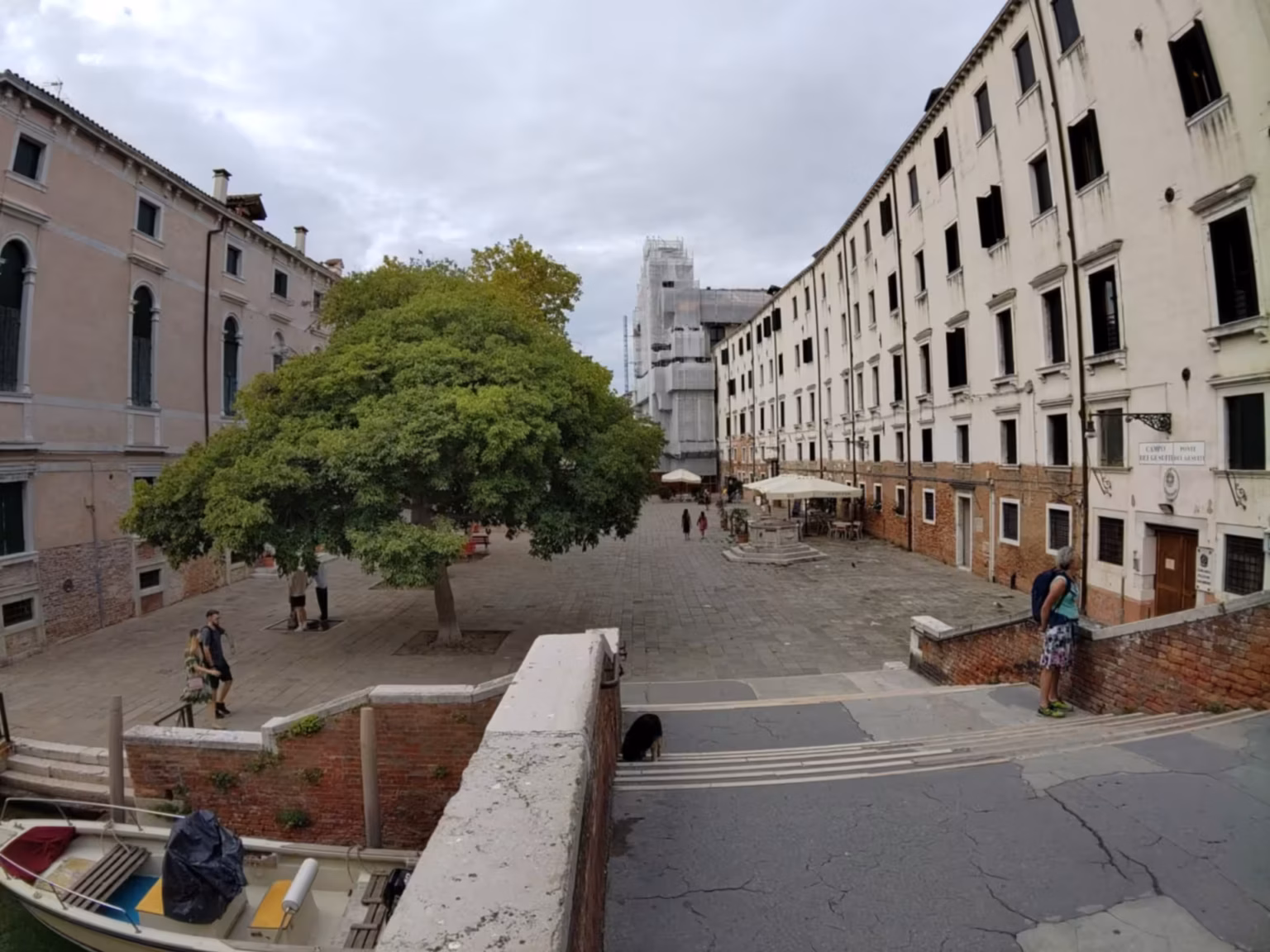 A photo of the Campo dei Gesuiti, wide angle, taken from above the side of the bridge. Much of the view is obstructed by a large tree, but the church can be seen in the background, under scaffolding, and the convent of the Jesuits on the right.