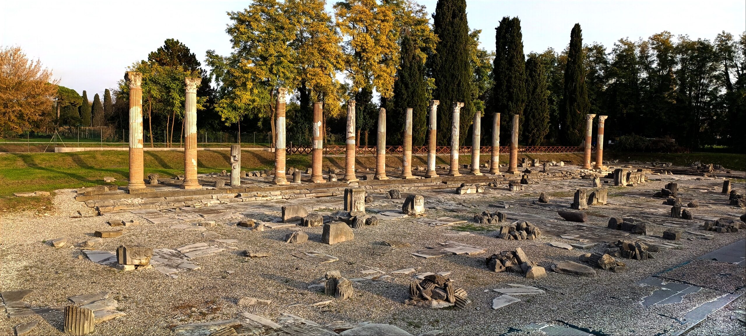 Ancient ruins. In the foreground an open area with broken columns and fragments of floor mosaics. In the background, a line of partially upright columns
