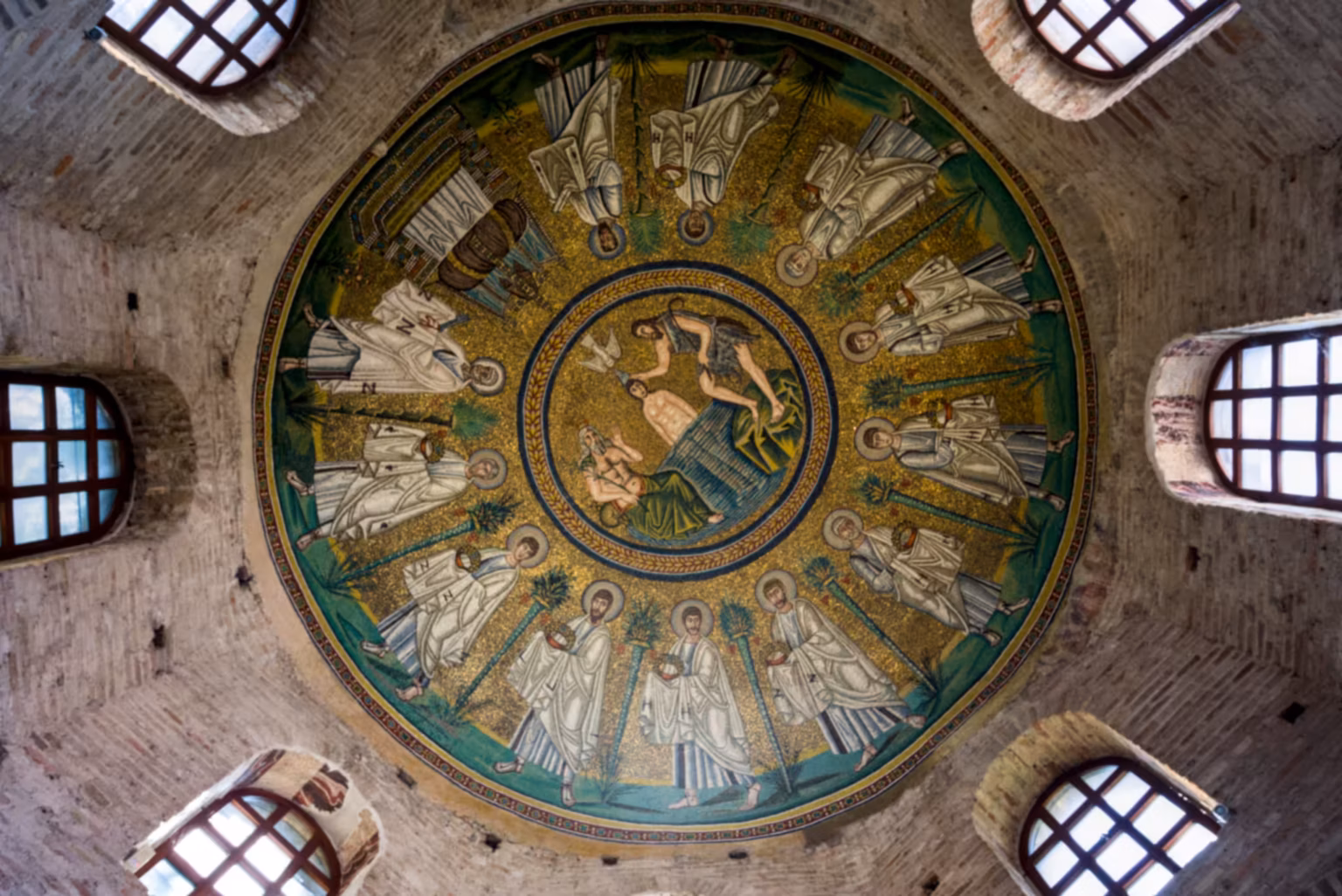 A view from below of the mosaics under the dome of the Arian baptistry in Ravenna, with a circle of Apostles, and an image of the baptism of Christ at the centre.